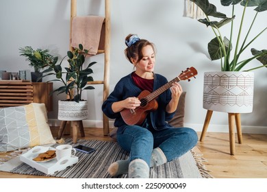 Young woman playing ukulele guitar while relaxing on floor cushion in modern scandinavian home interior.Musical Hobby, home fun concept. Resolution to learn new skills. Selective focus, - Shutterstock ID 2250900049