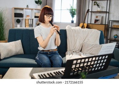 Young Woman Plays The Flute On Bright Living Room At Home. Girl Sitting On Sofa With A Musical Instrument Indoor. Pretty Young Woman Study To Play Flute.