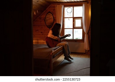 Young woman plays guitar and enjoys music alone, sitting by window of village house with cozy bed, wooden walls. Female in warm home clothes croon melody on musical instrument while rest on vacation. - Shutterstock ID 2255130723
