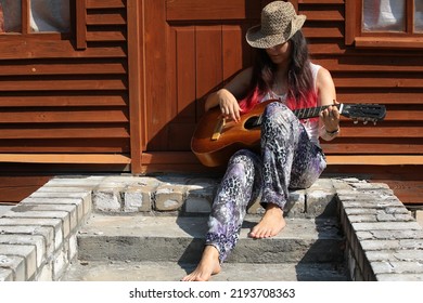 A Young Woman Plays The Guitar Sitting On The Steps Of A Wooden House With A Musical Instrument In A Hat With Long Hair Of An Unusual Red Color And Barefoot.Close-up Front View