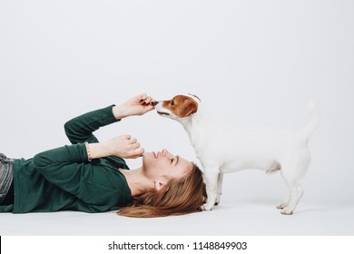 Young Woman Plays With Her Jack Russell Terrier Dog Isolated On White Background. Girl Lies Down And Looks Up On Her Dog. Owner And Dog Have Fun. Studio Portrait.