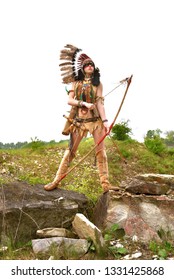 A young woman plays the part of a native American Indian. 
Dressed as a native Indian wearing a feathered headdress. 
She poses outdoors in a rocky nature surrounding.