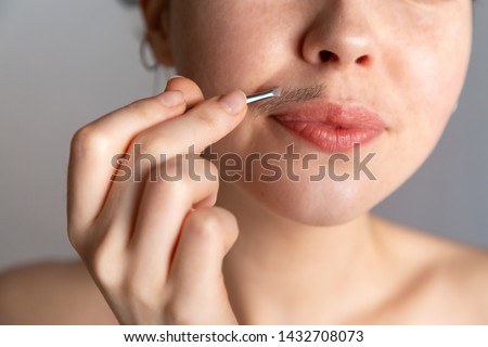 A young woman plucks her hair over her upper lip with tweezers. The concept of getting rid of unwanted facial hair
