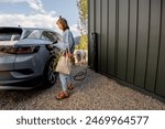 Young woman plugging a charger into electric vehicle near her house outdoors. Concept of modern technologies, EV cars and sustainable lifestyle