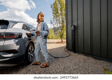 Young woman plugs a charger into electric vehicle standing with smart phone near her house outdoors. Concept of modern technologies, EV cars and sustainable lifestyle - Powered by Shutterstock