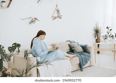 Young Woman Plus Size Model In A Blue Suit Works On A Laptop On The Couch In A Modern Light Interior With Home Garden And Macrame On The Wall