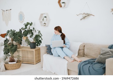 Young Woman Plus Size Model In A Blue Suit Works On A Laptop On The Couch In A Modern Light Interior With Home Garden And Macrame On The Wall