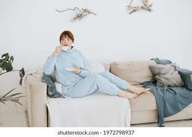 Young Woman Plus Size Model In A Blue Suit Drinking Coffee On The Couch In A Modern Light Interior With Home Garden And Macrame On The Wall