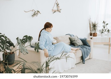 Young Woman Plus Size Model In A Blue Suit Drinking Coffee On The Couch In A Modern Light Interior With Home Garden And Macrame On The Wall