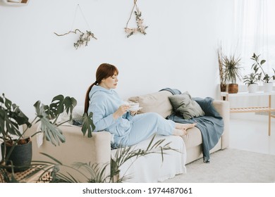 Young Woman Plus Size Model In A Blue Suit Drinking Coffee On The Couch In A Modern Light Interior With Home Garden And Macrame On The Wall