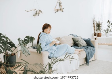 Young Woman Plus Size Model In A Blue Suit Drinking Coffee On The Couch In A Modern Light Interior With Home Garden And Macrame On The Wall