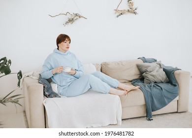 Young Woman Plus Size Model In A Blue Suit Drinking Coffee On The Couch In A Modern Light Interior With Home Garden And Macrame On The Wall