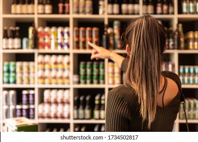 A Young Woman Pointing At Canned Beers In A Liquor Shop And Choosing Some. Beers From Trendy Small Breweries Displayed On Shelfs. Bokeh Background