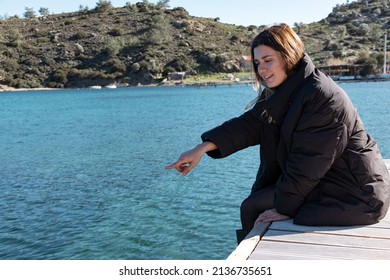 Young Woman Pointing Somewhere From Sea. She Is Showing Swimming Fish While Sitting On Dock. Winter Clothes. Happy And Smiling.	