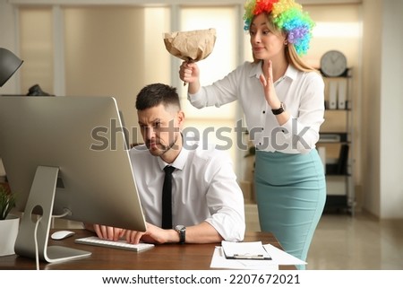 Young woman popping paper bag behind her colleague in office. Funny joke