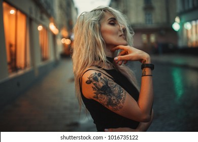 Young woman portrait with tattoo on shoulder standing on city street in evening. In background there are city lights. 