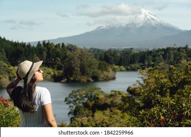 Young woman posing in profile and holding her hat with the view of Mount Taranaki behind. New Zealand.