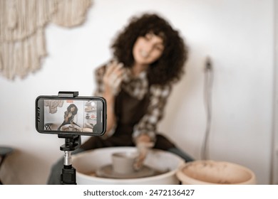 Young woman potter sculpting vase on potter's wheel while recording video blog with her smartphone. Creating pottery content for social media. - Powered by Shutterstock