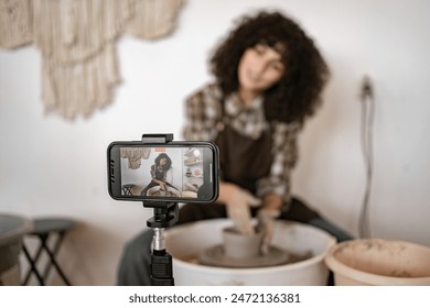 Young woman potter sculpts a jug using potter's wheel while recording video blog with smartphone. She appears focused and engaged in creative process. - Powered by Shutterstock