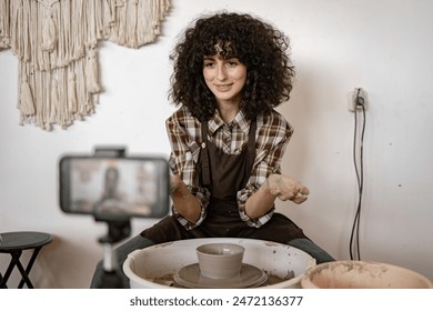 Young woman potter sculpts pottery using a potter's wheel while recording a video blog with her phone. Captures the creative process and artistry. - Powered by Shutterstock