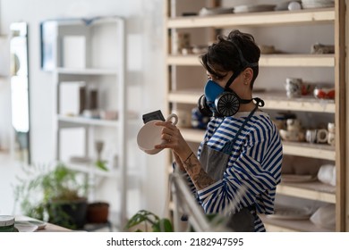 Young woman potter wearing respirator sanding ceramics with sandpaper or grinding block, female ceramist smoothing surface of pottery, creating handmade clay tableware to sell. Career in craft - Powered by Shutterstock