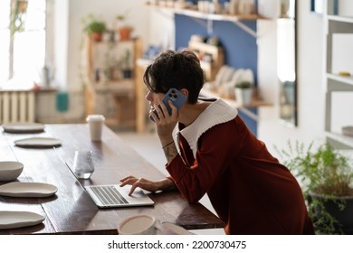 Young woman pottery business owner using laptop and talking with client by phone while working in workshop. Female artisan making money selling ceramics. Ceramic shop manager checking online order - Powered by Shutterstock