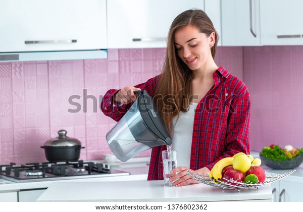Young\
woman pouring fresh filtered water from water filter into a glass\
for drink at kitchen. Water purification at home\
