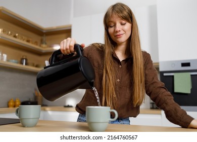 Young Woman Pouring Hot Water From Electric Kettle Into Mug In Kitchen. Girl Making Tea