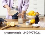 Young woman pouring lemon juice from jug into glass for preparing lavender lemonade at table in kitchen