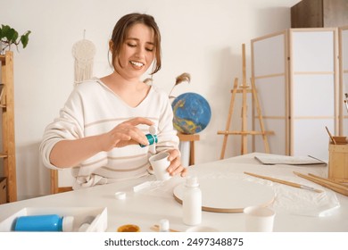 Young woman pouring paint into cup for fluid art at table in workshop - Powered by Shutterstock - Get 15% off with code: PIKWIZARD15