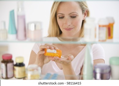 A Young Woman Pours Out Medicine Into Her Hand.  She Is Visible Through Her Medicine Cabinet.  Horizontal Shot.