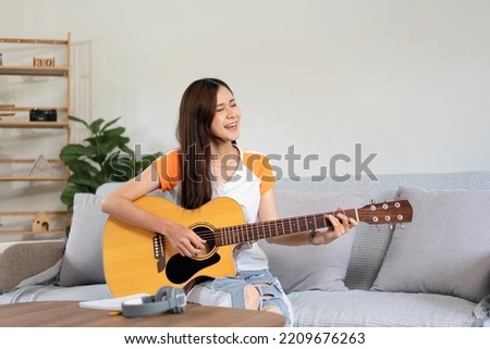 Young woman is practice basic guitar chords and playing guitar with pop songs while sitting on comfortable the couch in living room at home.