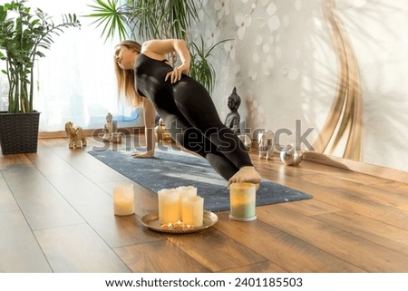 A young woman practices yoga and meditates in the Vasisthasana position surrounded by candles and green plants. Morning light. The concept of yoga classes at home. Online training concept.