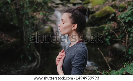 Young woman practicing breathing yoga pranayama outdoors in moss forest on background of waterfall. Unity with nature concept