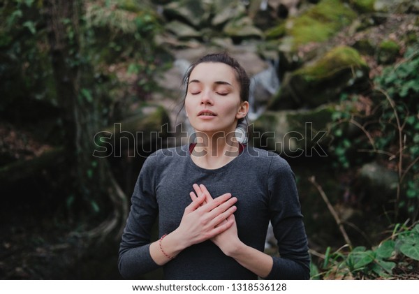 Young woman practicing breathing yoga pranayama\
outdoors in moss forest on background of waterfall. Unity with\
nature concept