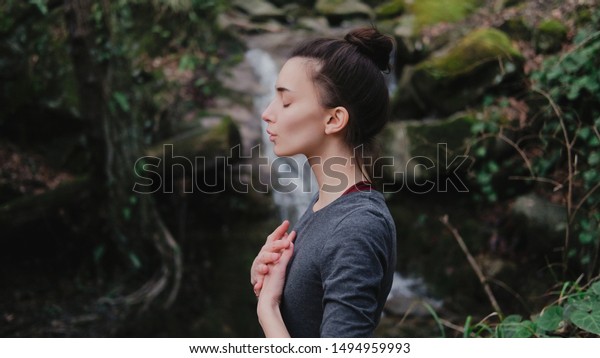 Young woman practicing breathing yoga pranayama\
outdoors in moss forest on background of waterfall. Unity with\
nature concept