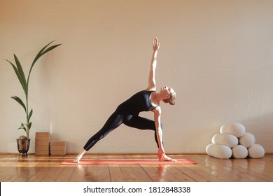 Young woman practicing the triangle Trikonasana yoga pose at home during self-isolation under covid-19 situation with head pillow under morning sunlight