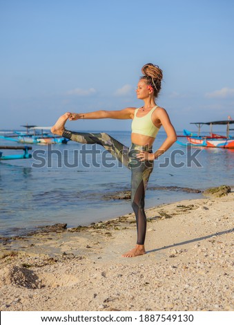 Young woman practicing Utthita Hasta Padangusthasana, Standing Big Toe Hold. Work out on the beach. Strong body. Yoga retreat. Balance and concentration. Thomas beach, Bali, Indonesia