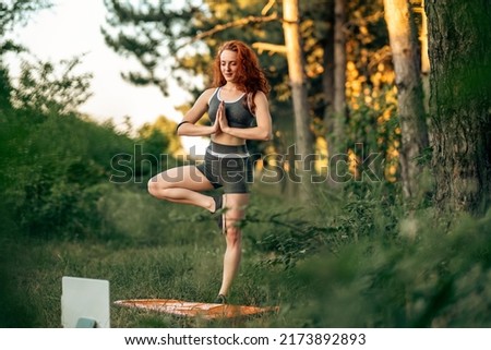 Young woman practicing yoga asana, balance, meditating while standing on one leg on sports mat on green grass in park. Using tablet for online class or virtual tutorials. Caucasian sportswoman.