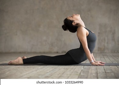 Молодая женщина, практикующая йогу, делает Кобра упражнения, Bhujangasana позы, тренировка, носить спортивную одежду, черные брюки и топ, крытый полный длины, серая стена в студии йоги