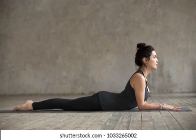 Young woman practicing yoga, doing Ardha bhudjangasana, Sphinx exercise, baby Cobra pose, working out, wearing sportswear, black pants and top, indoor full length, gray wall in yoga studio