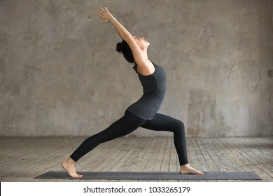 Young woman practicing yoga, doing Virabhadrasana 1 exercise, Warrior one pose, working out, wearing sportswear, black pants and top, indoor full length, gray wall in yoga studio