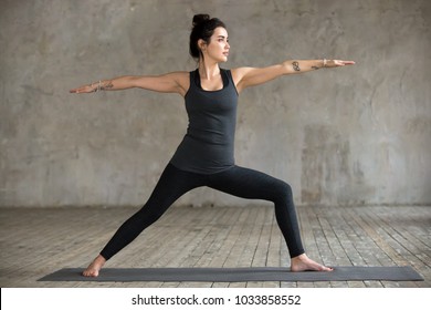 Young woman practicing yoga, doing Virabhadrasana 2 exercise, Warrior Two pose, working out, wearing sportswear, black pants and top, indoor full length, gray wall in yoga studio