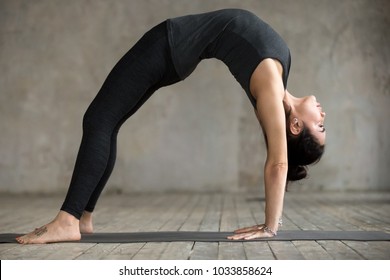 Young woman practicing yoga, doing Bridge exercise, Urdhva Dhanurasana pose, working out, wearing sportswear, black pants and top, indoor full length, gray wall in yoga studio