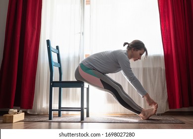 Young Woman Practicing Yoga, Doing Seated Forward Bend Pose, Paschimothanasana Or Caterpillar Yin Posture Using Chair At Home In The Living Room.