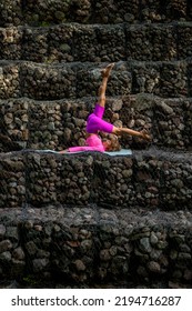 Young Woman Practicing Yoga Doing Halasana Exercise With One Leg Up. Standing In Plough Pose Against The Backdrop Of Rocky Steps. Side View.