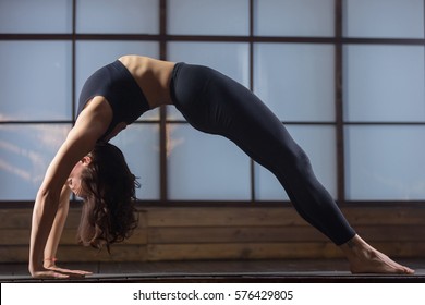 Young woman practicing yoga, doing Wild Thing, Flip-the-Dog pose, Camatkarasana, variation of Bending Side Plank exercise, Vasisthasana, working out, wearing sportswear, black tank top, pants 
