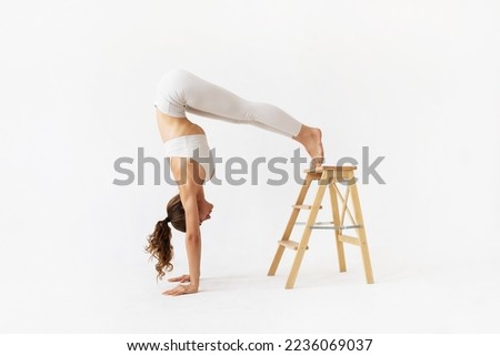 Young woman practicing yoga doing a variation of adho mukha vrikshasana exercise, head down tree pose, resting her feet on a wooden stand, exercising in white sportswear