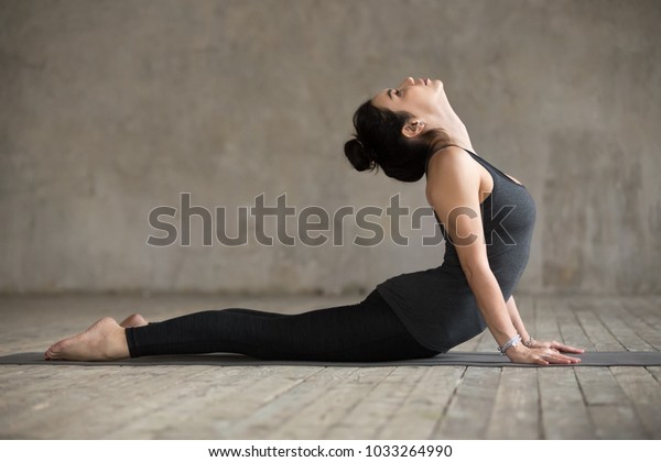 Young woman practicing yoga, doing Cobra exercise, Bhujangasana pose, working out, wearing sportswear, black pants and top, indoor full length, gray wall in yoga studio