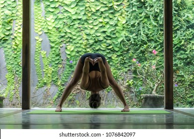young woman practicing yoga in Standing Forward Bend with Shoulder Opener pose (Uttanasana) in front of wall covered with green leaves